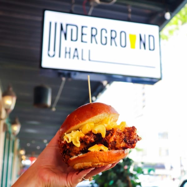Hand holding a SideChick HTX fried chicken sandwich with mac and cheese, in front of an "Underground Hall" sign.