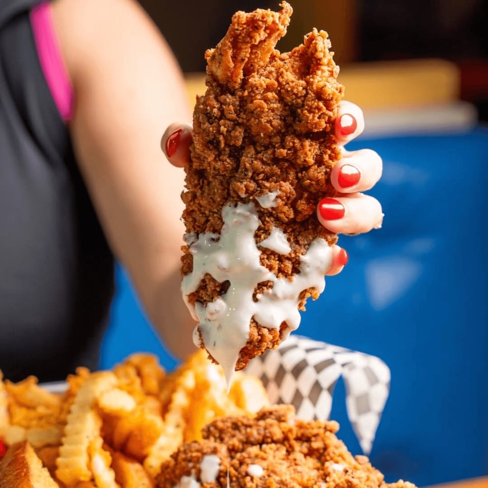 Hand holding a crispy fried chicken tender dripping with white sauce over a basket of fries.
